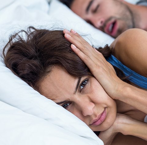 Woman in bed covering her ears next to a man sleeping with his mouth open